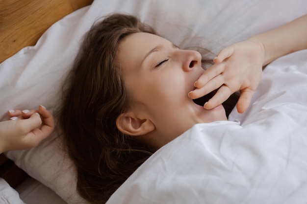 Une Jeune Fille Se Réveille Tôt Le Matin En Bâillant Couvrant Sa Bouche Avec Sa Main Dans Un Lit Avec Des Draps Blancs