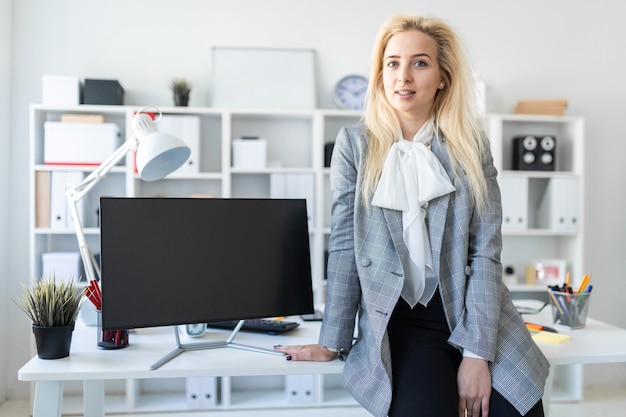 Jeune fille se tient dans le bureau, s'appuyant sur un bureau. A proximité se trouve un moniteur.