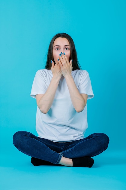 Jeune fille se tient la main sur la bouche en s'asseyant sur le sol sur fond bleu