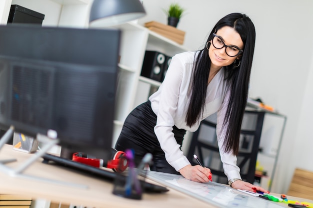 Une jeune fille se tient près d'un bureau d'ordinateur et dessine un marqueur sur un tableau magnétique.