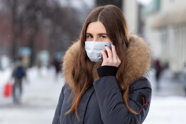 Une jeune fille se tient près de la route et parle sur le smartphone dans un masque médical Protection contre les épidémies de virus grippe et pollution de l'air par les voitures de la ville