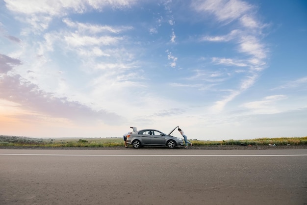 Une jeune fille se tient près d'une voiture en panne au milieu de l'autoroute au coucher du soleil et essaie d'appeler à l'aide par téléphone. Dépannage et réparation de la voiture. En attente d'aide.