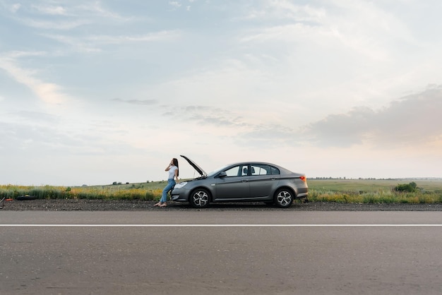 Une jeune fille se tient près d'une voiture en panne au milieu de l'autoroute pendant le coucher du soleil et essaie d'appeler de l'aide au téléphone En attente d'aide Service de voiture Panne de voiture sur la route