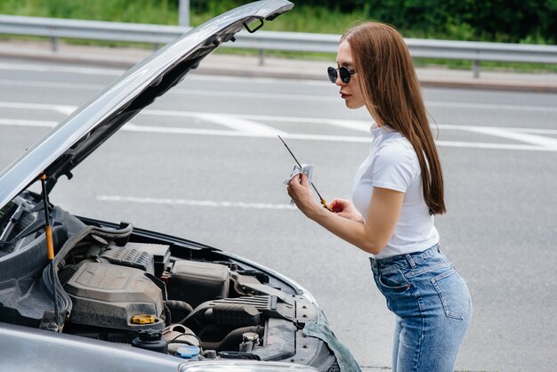 Une jeune fille se tient près d'une voiture en panne au milieu de l'autoroute et vérifie le niveau d'huile dans le moteur. Panne et panne de la voiture. En attente d'aide.