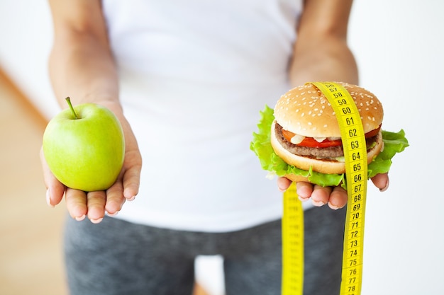 Une jeune fille séduisante fait le choix entre des aliments sains et nocifs tenant une pomme et un hamburger dans ses mains.