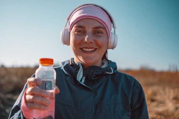 Jeune fille souriante active tenant une bouteille d'eau propre à la main tout en se tenant dans la nature après avoir fait de l'exercice à l'extérieur