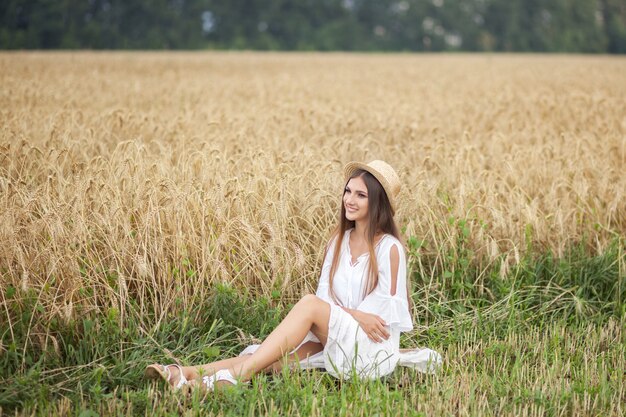 Jeune fille souriante au chapeau de paille et en robe blanche appréciant la nature du champ de blé. vacances d'été