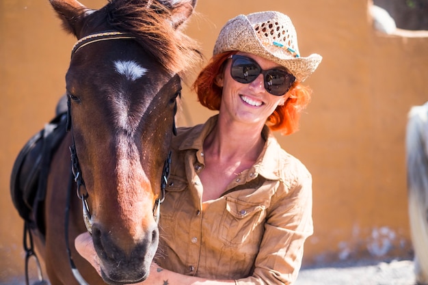 Jeune fille souriante aux cheveux roux et chapeau de cow-boy posant avec son cheval.