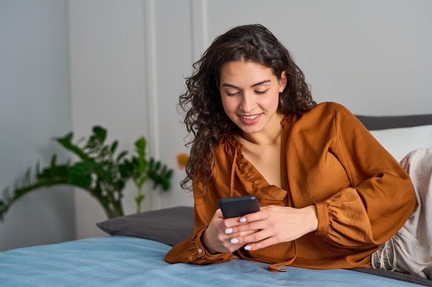 Jeune fille souriante en blouse de soie de couleur bronze allongée sur un lit double et textos