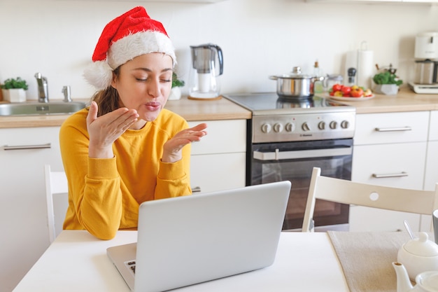 Jeune fille souriante en bonnet de Noel ayant un chat de réunion virtuelle en vacances assis sur la cuisine à la maison