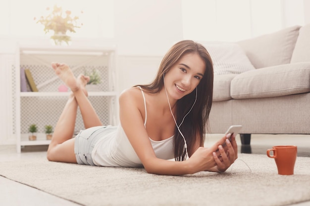Jeune fille souriante écoutant de la musique en ligne sur un smartphone dans des écouteurs, allongée sur le sol avec du café à la maison, espace de copie