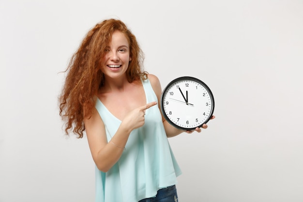 Jeune fille souriante jolie femme rousse dans des vêtements légers décontractés posant isolé sur fond de mur blanc, portrait en studio. Concept de mode de vie des gens. Maquette de l'espace de copie. Pointer l'index sur l'horloge.