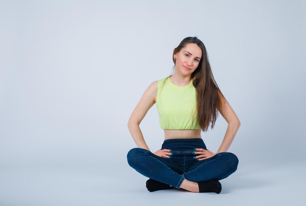 Jeune fille souriante met les mains sur la taille en s'asseyant sur le sol sur fond blanc