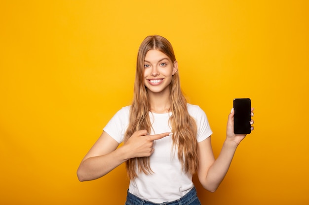jeune fille souriante, pointant du doigt au smartphone isolé sur jaune
