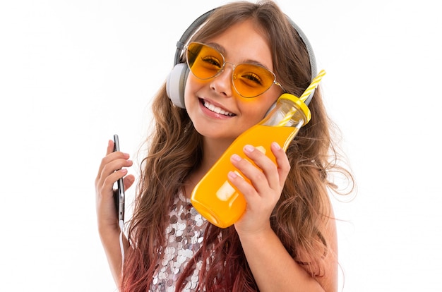 Photo jeune fille souriante portant une robe pailletée et des lunettes de soleil jaune vif, avec de gros écouteurs blancs, écoutant de la musique et tenant un smartphone mince et une bouteille avec du jus d'orange isolé