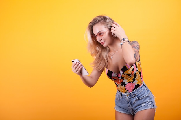 Jeune fille souriante regardant l'écran du téléphone dans ses mains avec des lunettes de soleil en studio sur fond jaune