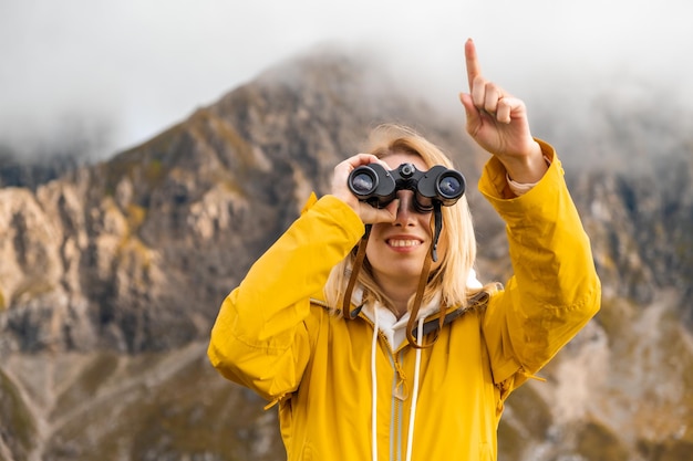 Jeune fille souriante regardant à travers des jumelles et montrant du doigt au loin avec des montagnes incroyables