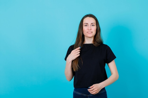 Une jeune fille souriante regarde la caméra en tenant ses cheveux sur fond bleu