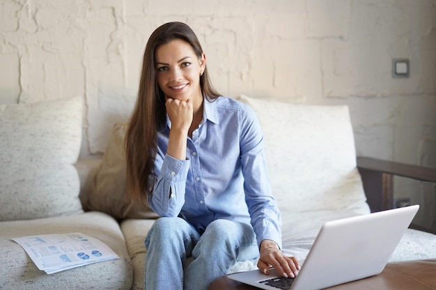 Une jeune fille souriante s'assoit sur un canapé en regardant un webinaire sur un ordinateur portable