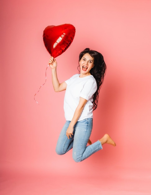 Jeune fille souriante sautant avec un ballon en forme de coeur rouge sur fond rose. La Saint-Valentin