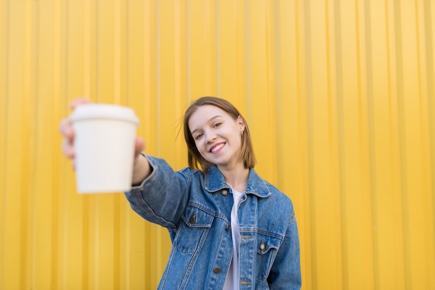 jeune fille souriante se dresse sur fond jaune et offre une tasse de café en papier.