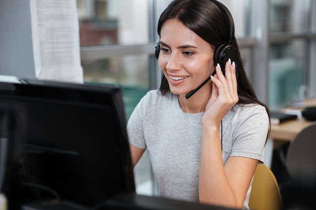 Jeune fille souriante en t-shirt assis près de l'ordinateur dans un casque au bureau. Centre d'appel