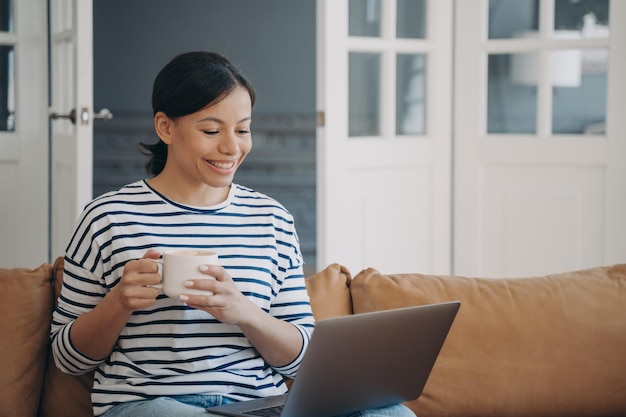 Jeune fille souriante avec une tasse de café en regardant un film ou en communiquant par appel vidéo sur un ordinateur portable sur un canapé à la maison