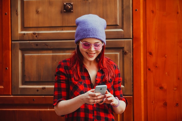 Jeune fille souriante utilisant un téléphone intelligent sur un mur en bois. Fille hipster élégante aux cheveux rouges et pirsing sur les lèvres