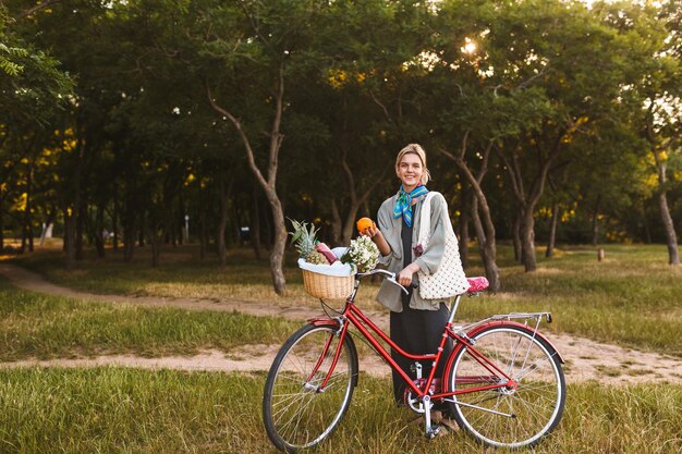 Jeune fille souriante avec vélo rouge et panier plein de fleurs sauvages et de fruits regardant joyeusement à huis clos tenant orange à la main dans le parc