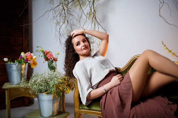 La jeune fille sourit dans une jupe marron est assise sur le canapé. autour sont des fleurs fraîches dans un vase.