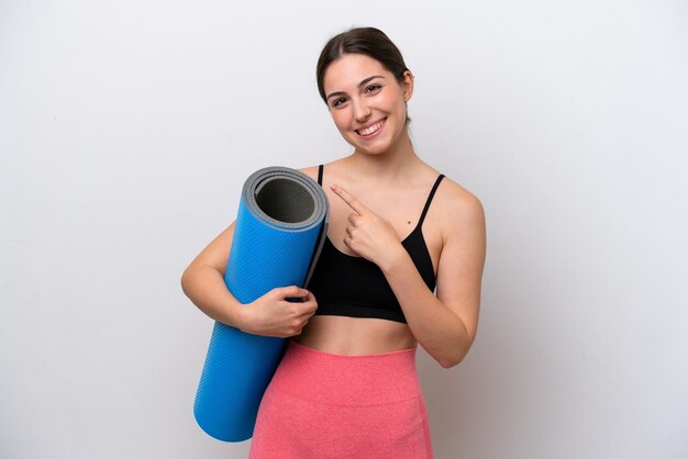 Jeune fille sportive allant à des cours de yoga tout en tenant un tapis isolé sur fond blanc pointant vers le côté pour présenter un produit