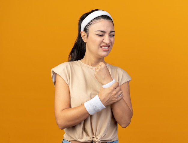 Jeune fille sportive caucasienne douloureuse portant un bandeau et des bracelets tenant le poignet avec les yeux fermés isolé sur un mur orange avec espace de copie