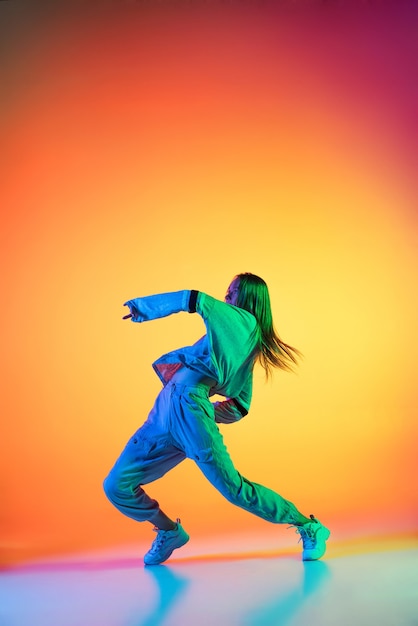 Jeune fille sportive dansant le hip-hop dans des vêtements élégants sur fond coloré à la salle de danse en néon. Culture jeunesse, mouvement, style et mode, action.