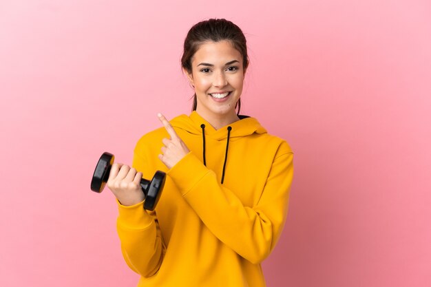 Jeune fille sportive faisant de l'haltérophilie sur fond rose isolé pointant vers le côté pour présenter un produit