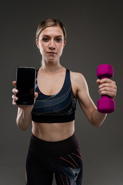 Jeune fille sportive avec de longs cheveux blonds farcis en queue, belle apparence, corps de sport, en haut noir et legines, tient des gants en caoutchouc violet et un téléphone dans ses mains