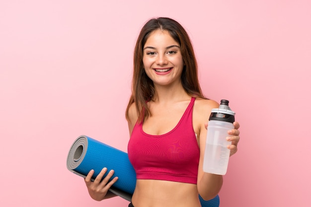 Jeune fille sportive sur mur rose isolé avec une bouteille d'eau de sport et un tapis