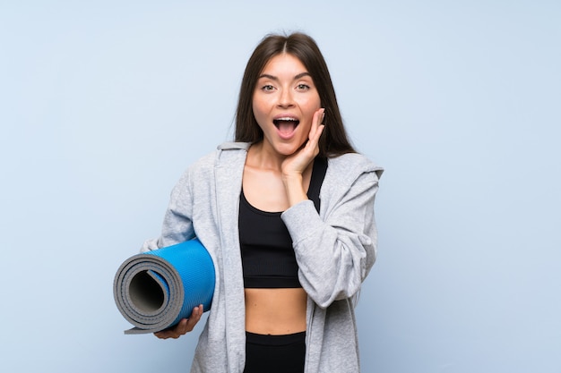 Jeune fille sportive avec tapis sur mur bleu isolé avec une expression faciale surprise et choquée