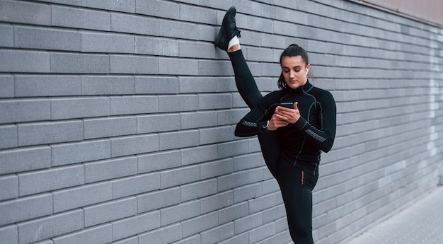 Jeune fille sportive avec téléphone en tenue de sport noire faisant des jambes qui s'étendent à l'extérieur en utilisant un mur gris