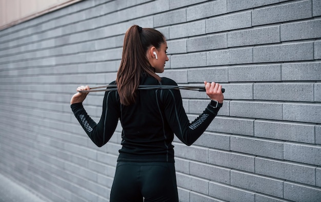 Jeune fille sportive en tenue de sport noire debout avec une corde à sauter dans les mains à l'extérieur près d'un mur gris