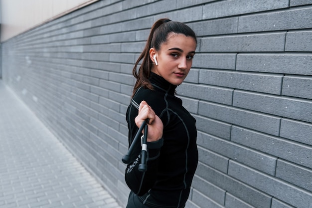 Jeune fille sportive en tenue de sport noire debout avec une corde à sauter dans les mains à l'extérieur près d'un mur gris