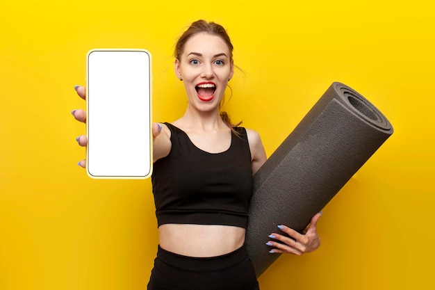 une jeune fille sportive en tenue de sport avec un tapis de yoga montre un écran de smartphone vierge sur fond jaune