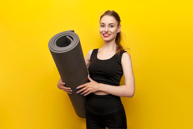 jeune fille sportive en tenue de sport avec tapis de yoga sourit sur fond jaune isolé