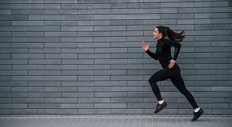 Jeune fille sportive en vêtements de sport noirs courant à l'extérieur près du mur gris