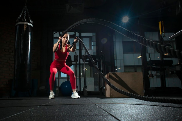 Jeune fille sportive en vêtements de sport rouges s'entraîne avec des cordes dans une femme motivée par une salle de sport noire et foncée