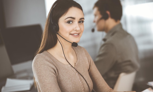 Une jeune fille sympathique dans des casques parle au client d'une entreprise, alors qu'elle est assise au bureau dans un bureau moderne avec son collègue. Opérateurs de centre d'appels au travail.