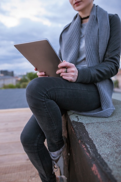 Jeune fille avec tablette travaillant sur fond de ciel nuageux. Femme intelligente assise sur le toit du bâtiment en ville pendant le travail. Blogueur, travail à distance, concept internet sans fil