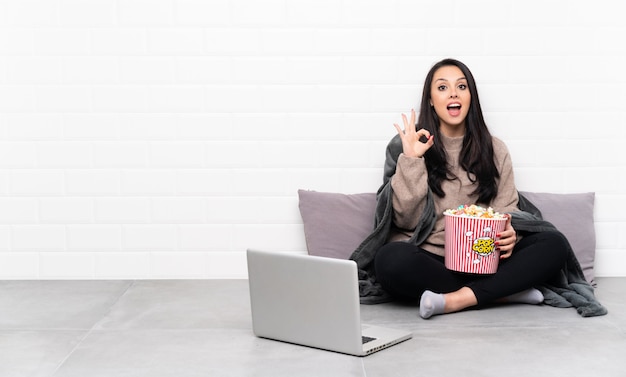 Jeune fille tenant un bol de pop-corn et montrant un film dans un ordinateur portable montrant le signe ok et le geste du pouce vers le haut