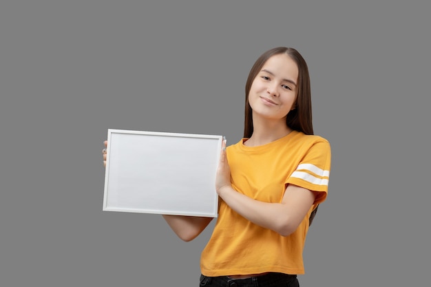 Jeune fille tenant un cadre blanc horizontal avec une maquette dans ses mains, souriante, isolée sur fond gris