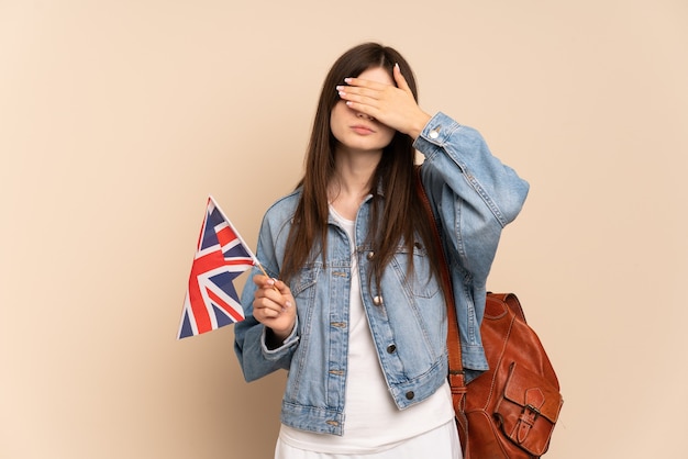 Jeune fille tenant un drapeau du Royaume-Uni isolé sur beige couvrant les yeux par les mains