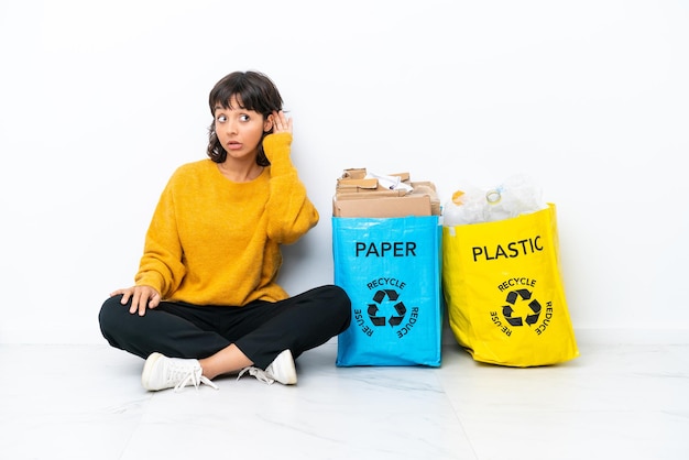 Jeune fille tenant un sac plein de plastique et de papier assis sur le sol isolé sur fond blanc écoutant quelque chose en mettant la main sur l'oreille